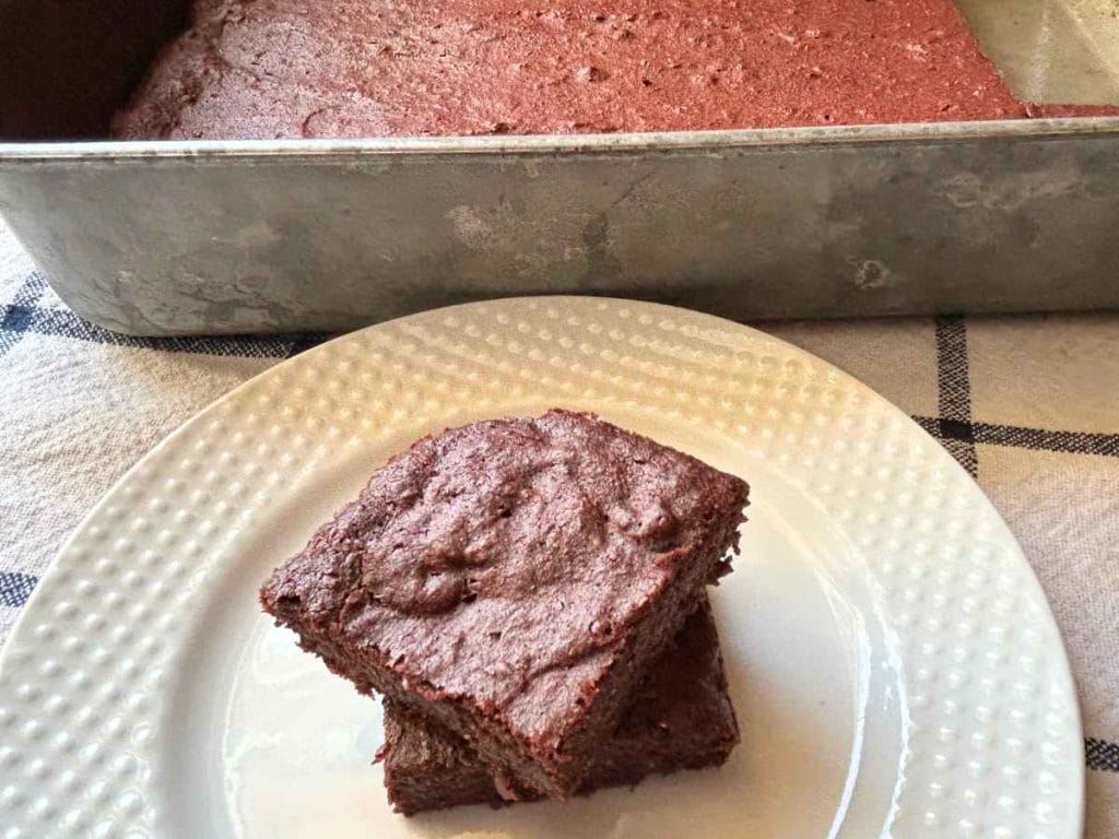 two beet brownies stacked on a white plate near pan of brownies