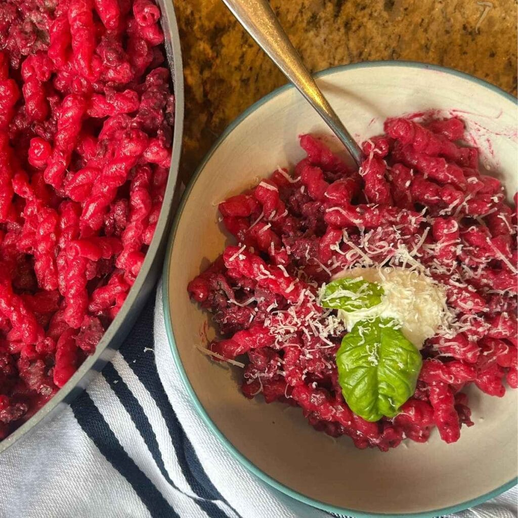 skillet full of bright pink noodles with a sprig of basil in the center next to a serving of the beet pasta in a bowl