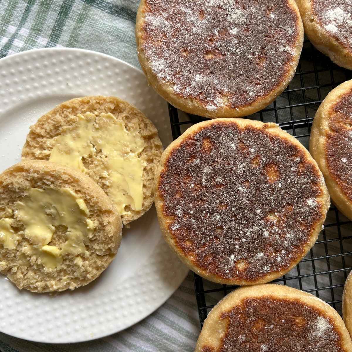 close up of English muffins on a cooling rack next to a split and buttered one on a white plate