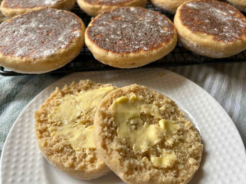 close up of English muffins on a cooling rack behind a split and buttered one on a white plate