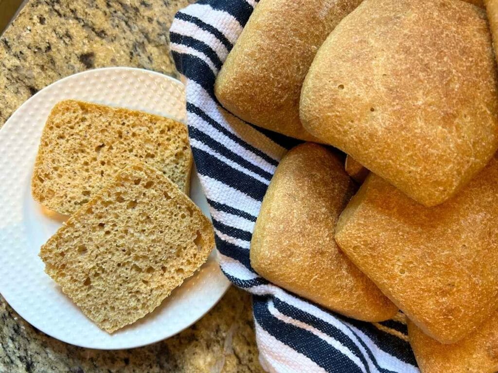 a sliced ciabatta roll on a white plate next to towel lined basket of whole rolls