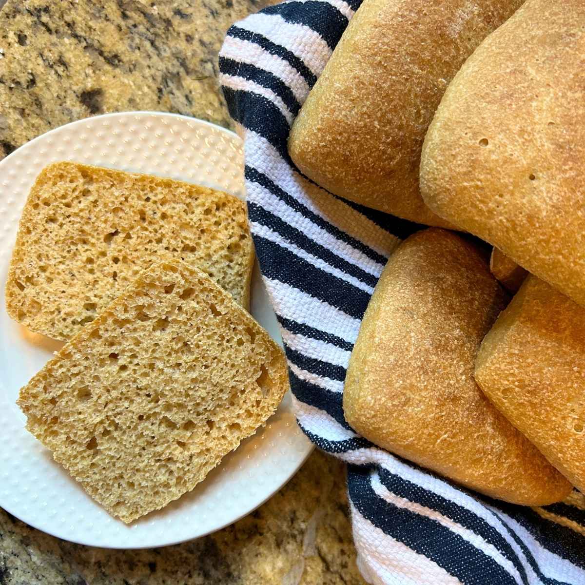 a sliced ciabatta roll on a white plate next to towel lined basket of whole rolls