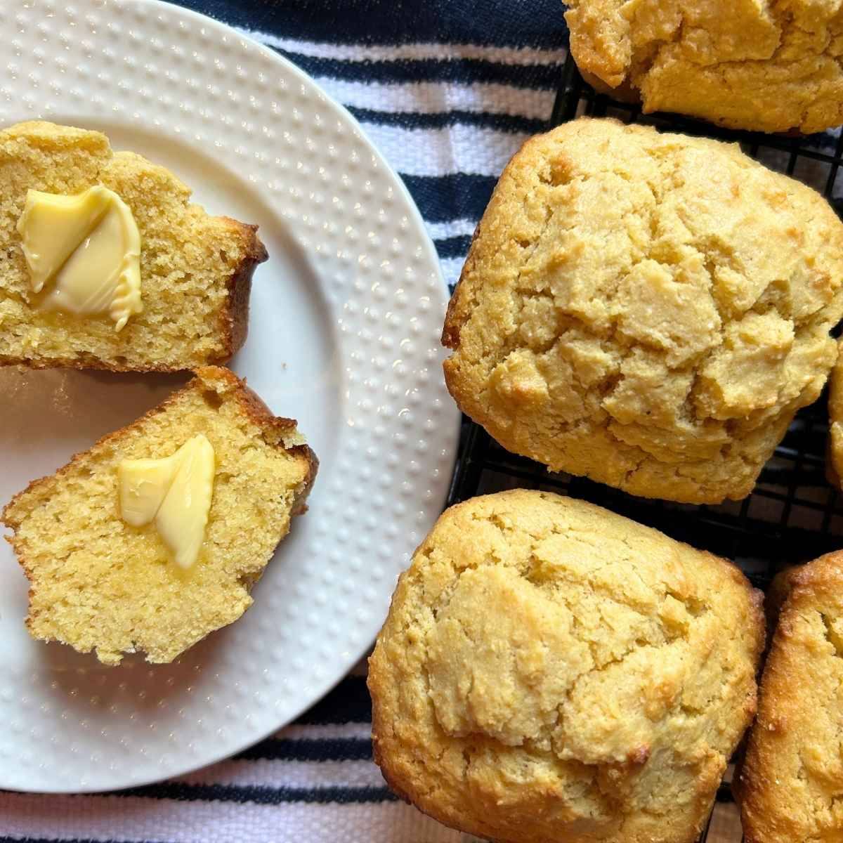 a halved cornbread muffin with butter pats on each side on a plate next to a few whole muffins