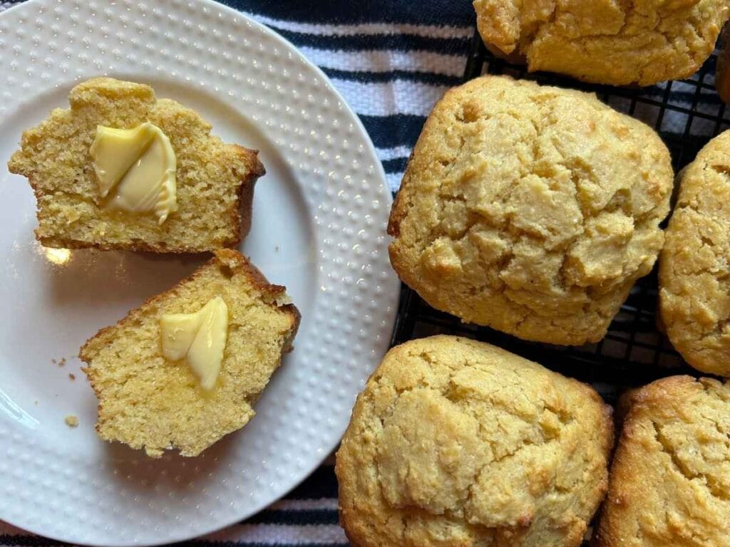 a halved cornbread muffin with butter pats on each side on a plate next to a few whole muffins