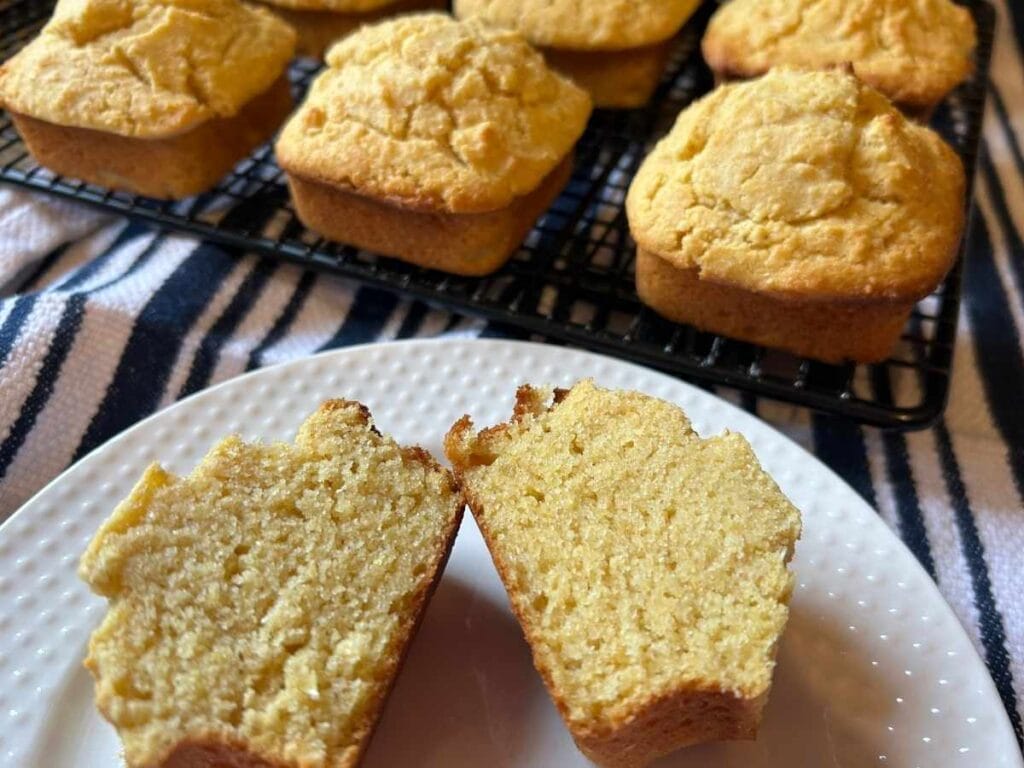 a halved cornbread muffin on a plate in front of a few whole muffins