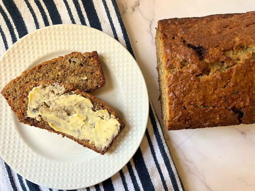 two slices of banana bread, one is buttered, on a white plate next to the rest of the loaf