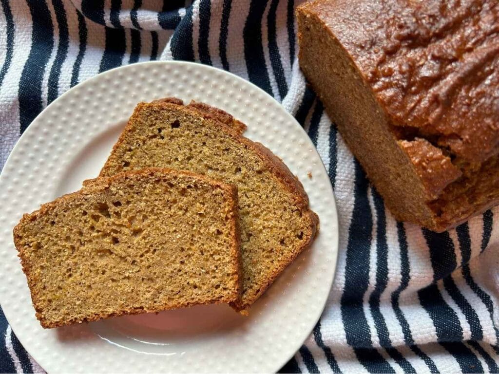 two slices of sourdough discard pumpkin bread on a white plate next to the rest of the loaf on a blue striped towel