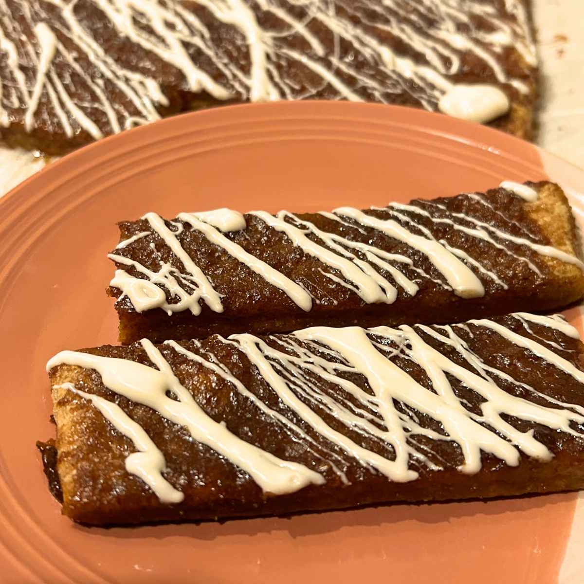 two sourdough pumpkin sticks drizzled with icing on an orange plate in front of the rest of the batch