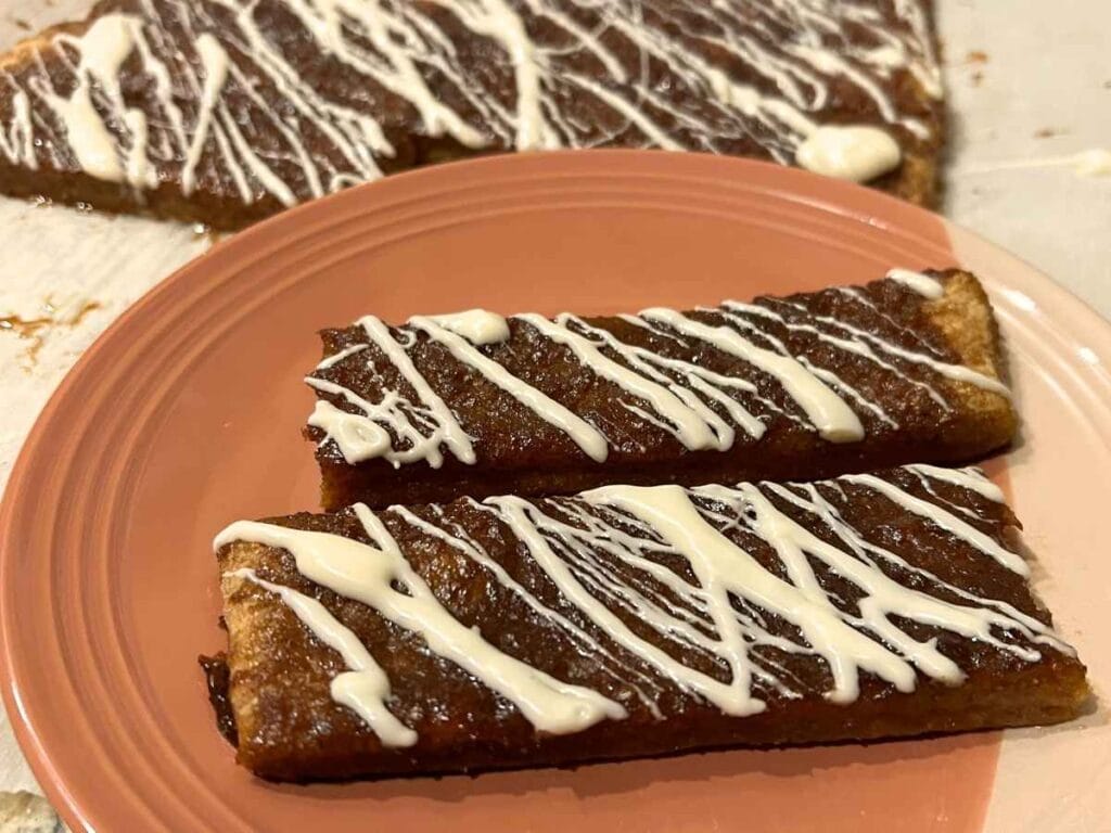 two sourdough pumpkin sticks drizzled with icing on an orange plate in front of the rest of the batch