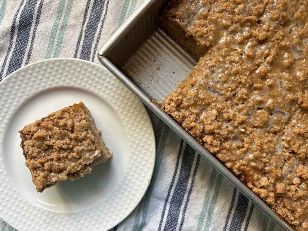top view of a slice of coffee cake on a white plate next to the pan with the rest of the cake