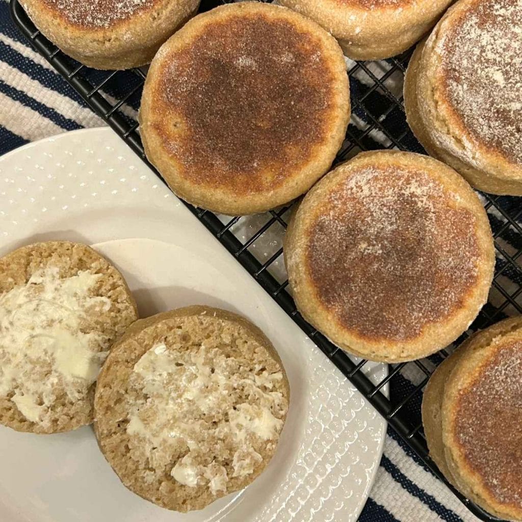 top view of an english muffin split and buttered on a white plate next to a cooling rack with the rest of the batch