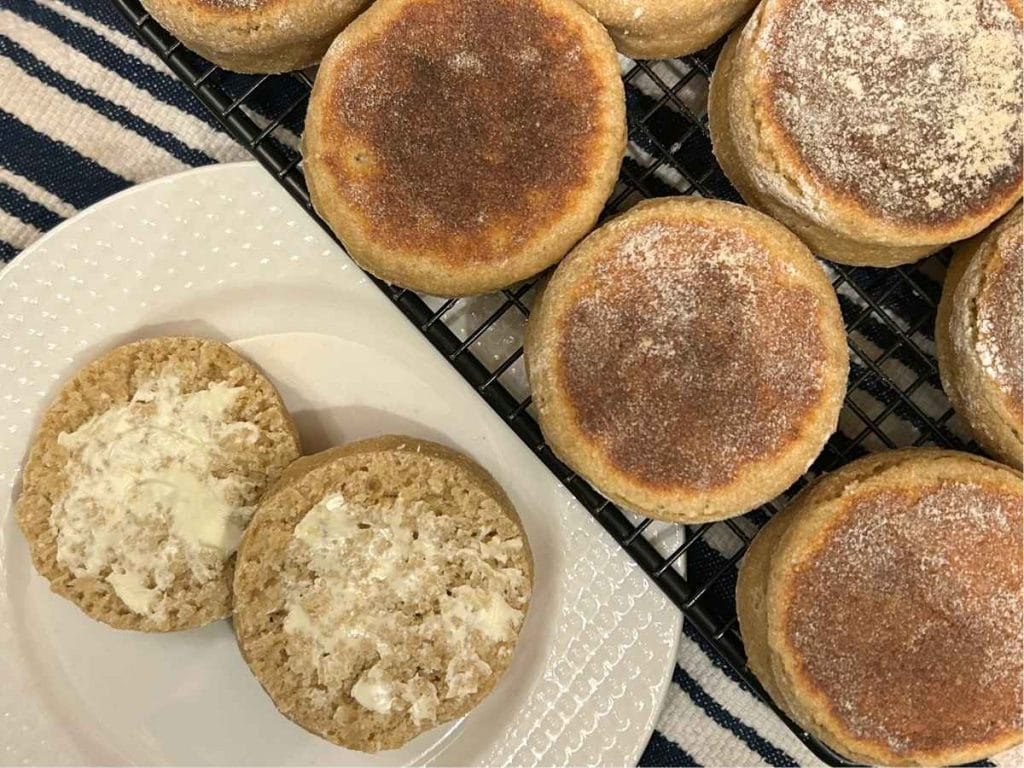 top view of an english muffin split and buttered on a white plate next to a cooling rack with the rest of the batch