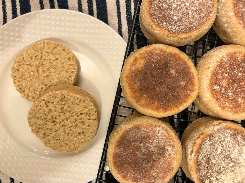 top view of an english muffin split on a white plate next to a cooling rack with the rest of the batch