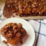 a sourdough pecan sticky bun on a white plate in front of the pan of the rest of the buns