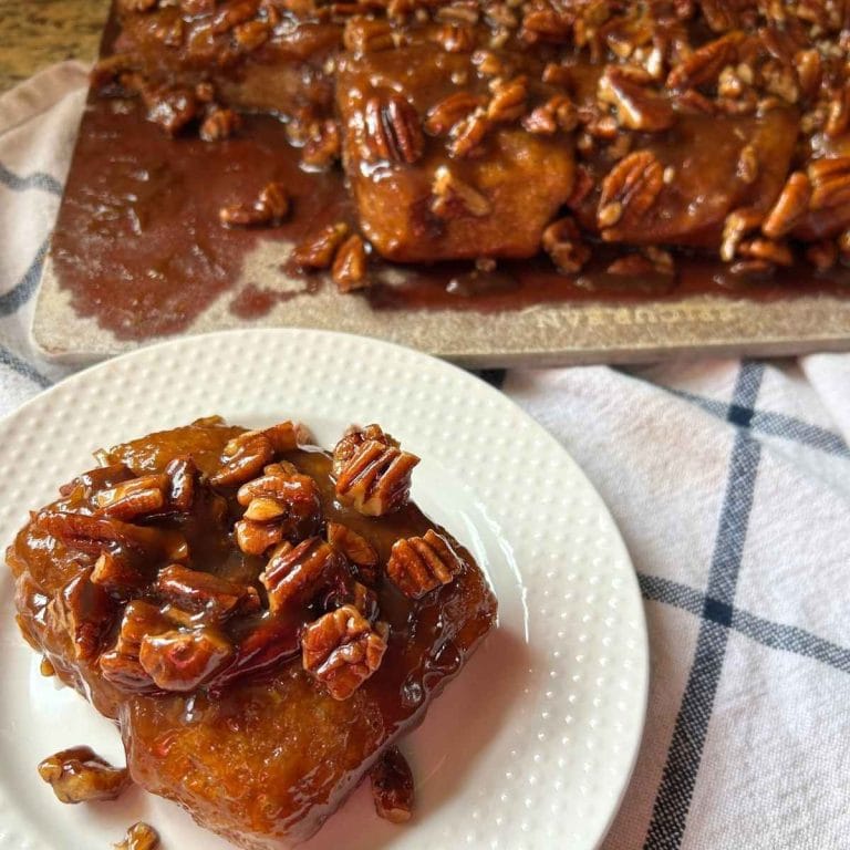 a sourdough pecan sticky bun on a white plate in front of the pan of the rest of the buns
