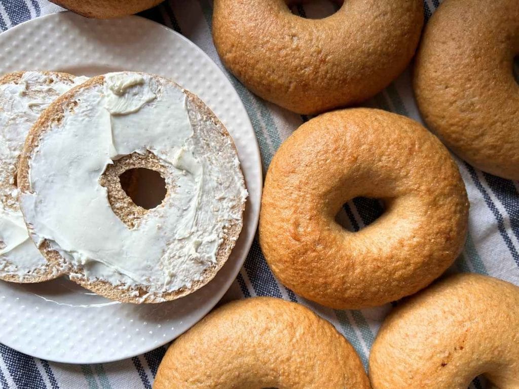 a cut bagel with cream cheese on a plate next to the rest of the batch of sourdough bagels on a striped kitchen towel