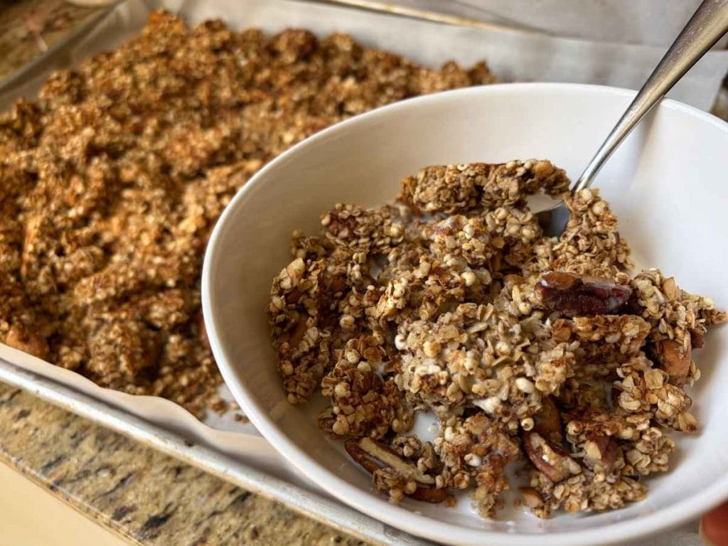 homemade sourdough maple nut granola in a bowl with milk next to the pan with the rest of the batch