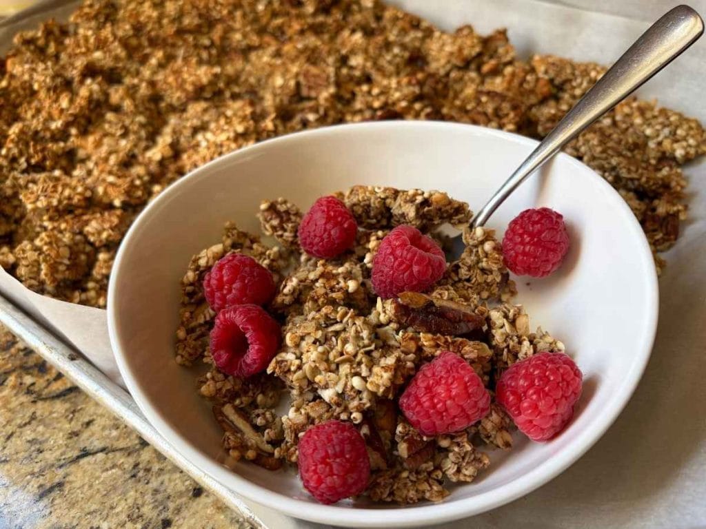 homemade sourdough maple nut granola in a bowl with raspberries next to the pan with the rest of the batch