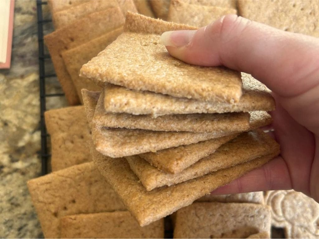 a hand holding a stack of graham crackers over a cooling rack filled with the rest