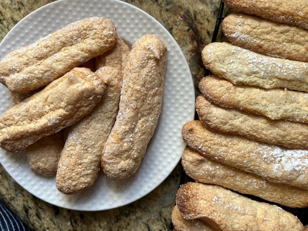 baked ladyfingers arranged on a plate next to some on a cooling rack