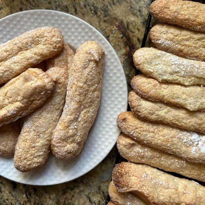 baked ladyfingers arranged on a plate next to some on a cooling rack