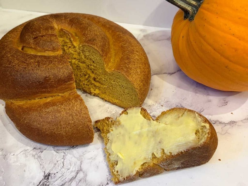 a slice of buttered pumpkin challah bread next to the loaf and a pumpkin
