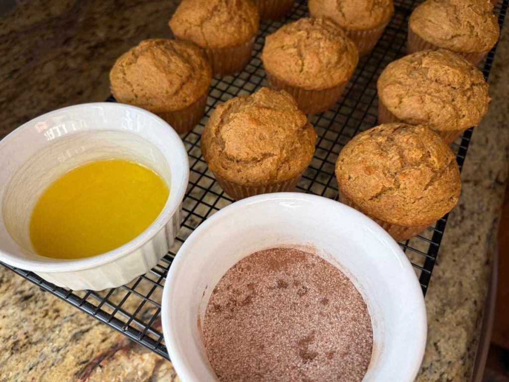 baked pumpkin muffins next to a bowl of melted butter and a bowl of cinnamon sugar