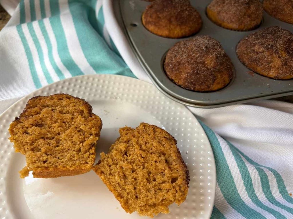 a pumpkin spice muffin halved on a white plate near the muffin pan with the rest of the batch